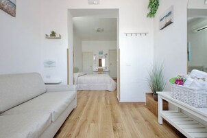 Egy modern, minimalista stílusú szoba, melyben fa padló, fehér kanapé, ágy, egy kis asztal és egy falra szerelt polc található, amin egy növény áll.