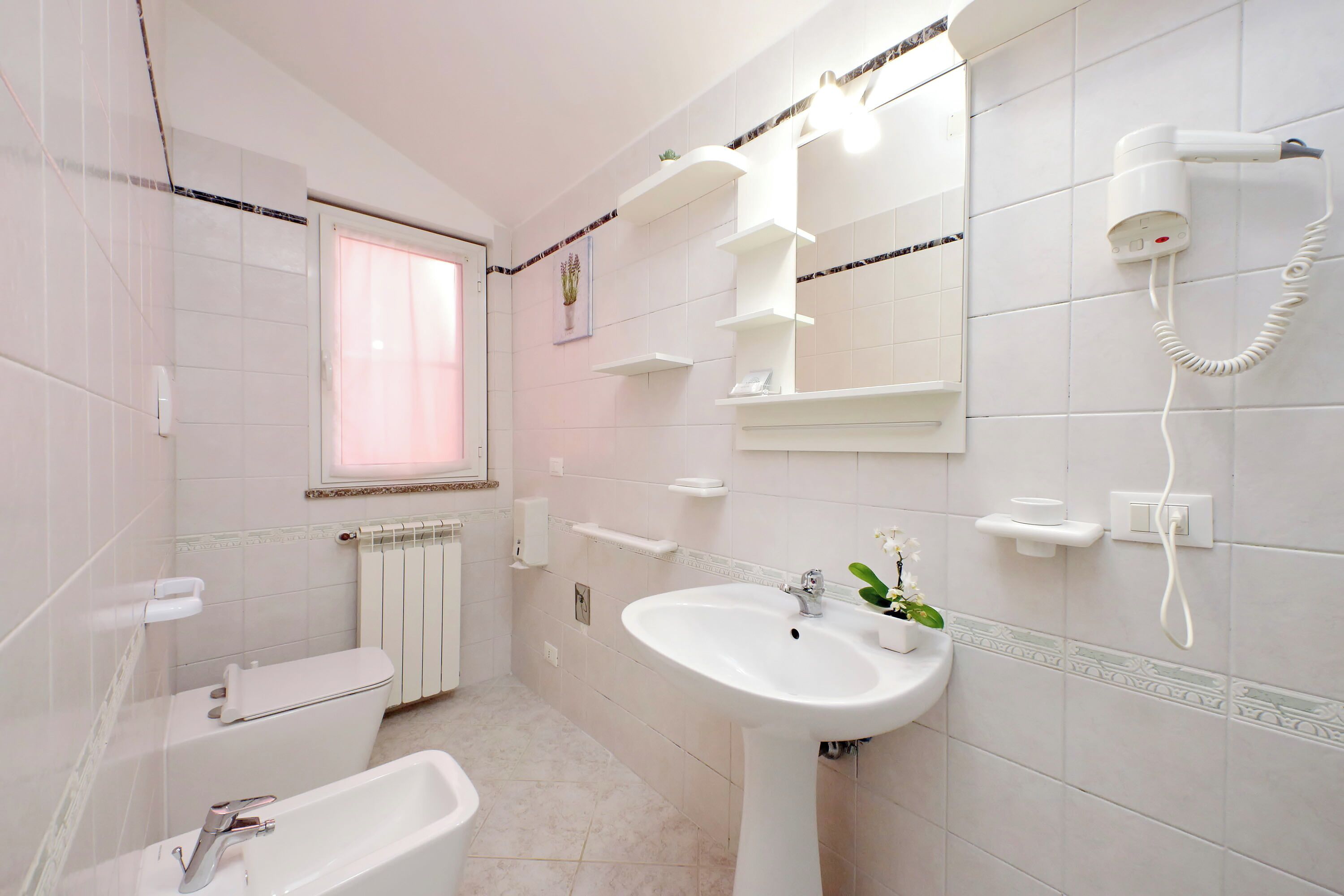 Une salle de bain équipée de toilettes, d’un lavabo et d’une baignoire. Il y a des étagères au-dessus du lavabo et un sèche-cheveux fixé au mur.