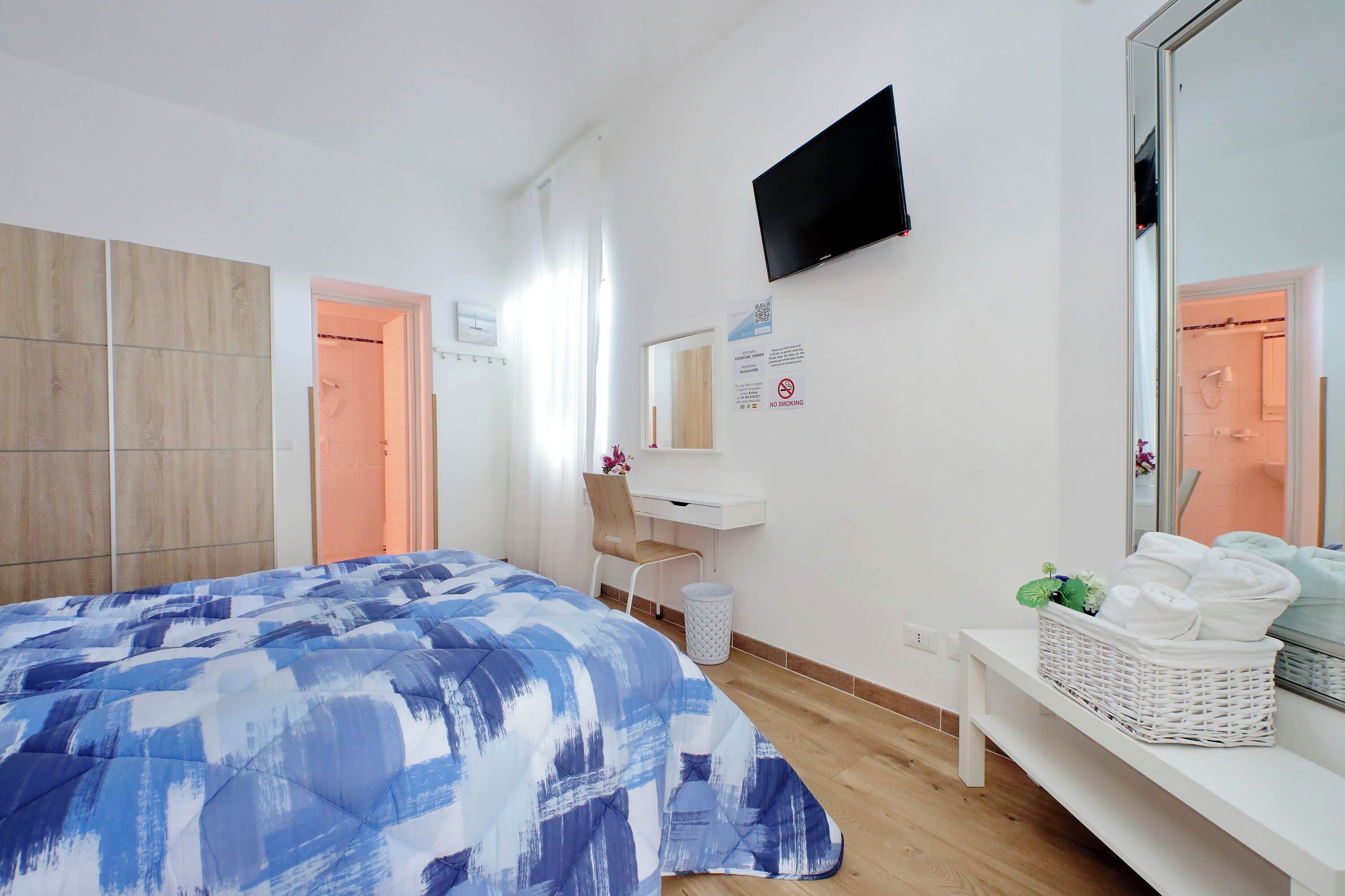 Quarto de hotel com uma cama, uma escrivaninha, uma televisão, um espelho e uma cesta de toalhas.