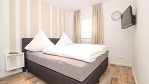 Ein Schlafzimmer mit einem Bett, einem Nachttisch, einem Wand-Fernseher und einem Fenster mit Vorhängen.