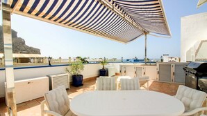 A rooftop patio with a round table, white cushioned chairs, a striped umbrella, potted plants, and a grill. - Playa Azul , Luxury Penthouse With Spectacular Roof Terrace (Mogan)