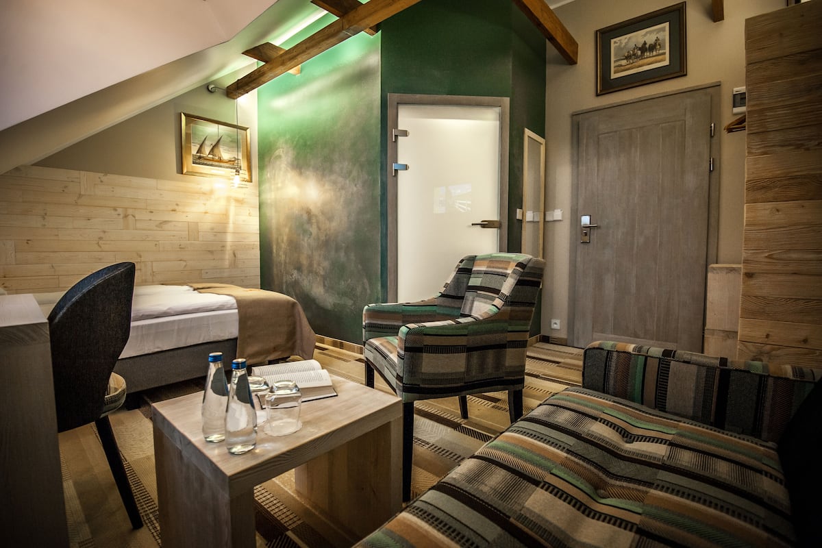 Een knus ingerichte kamer met een bed, een stoel, een bank en een klein tafeltje met een boek en flessen.