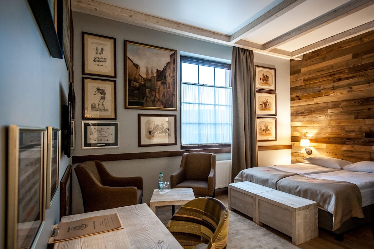 Hotelkamer met houten hoofdeinde, groot raam met gordijnen, bureau met stoel en bed met nachtkastje.