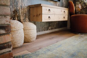 Une commode en bois avec des tiroirs, deux poufs en fibres tressées et une chaise rouge dans une pièce dotée d’un plancher en bois et d’un mur à texture.