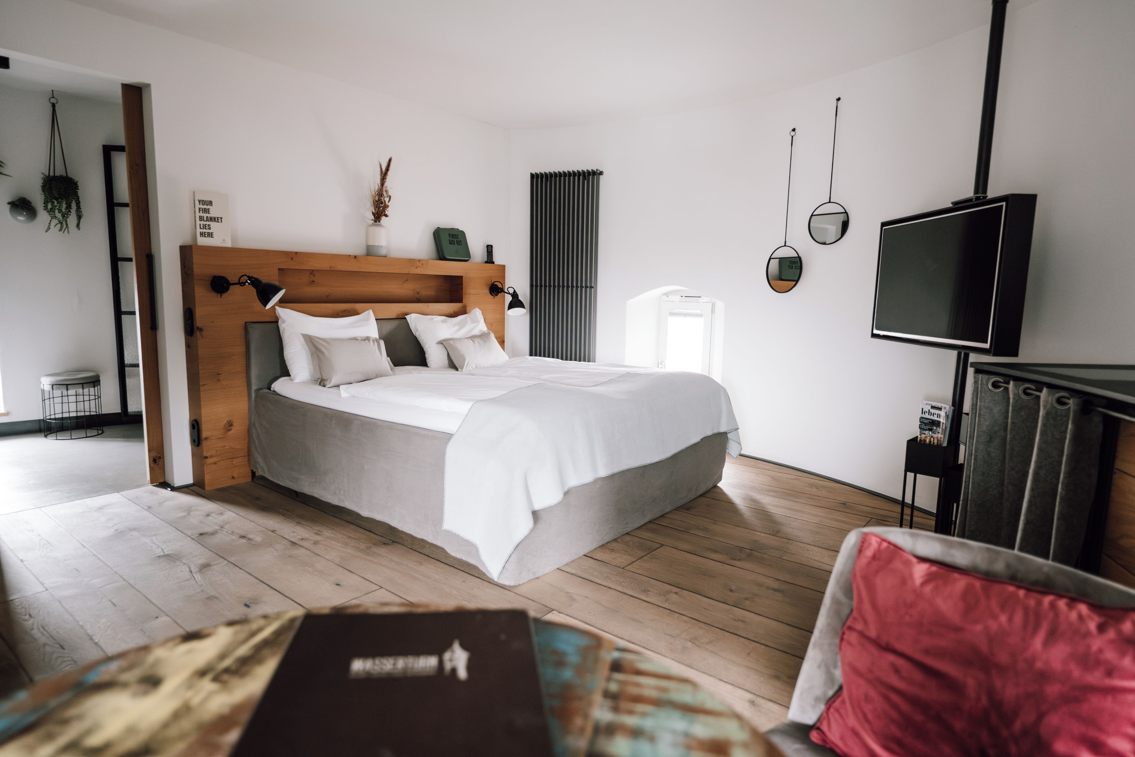 A modern bedroom with a wooden headboard, a bed with white bedding, a television, and a small table with books.