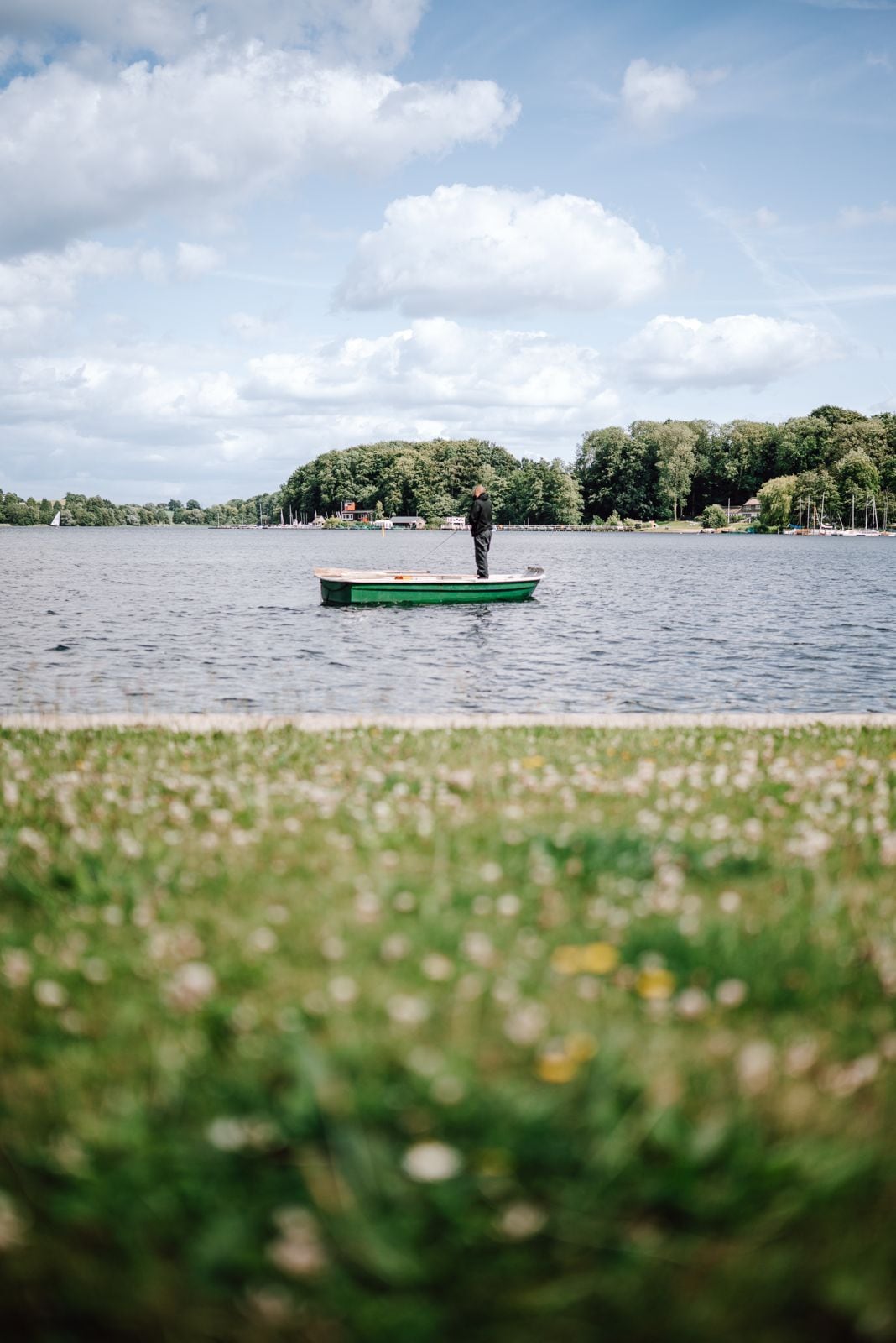 Une personne est en train de pêcher depuis un petit bateau vert sur un lac calme, avec un paysage verdoyant au premier plan et des arbres en arrière-plan.