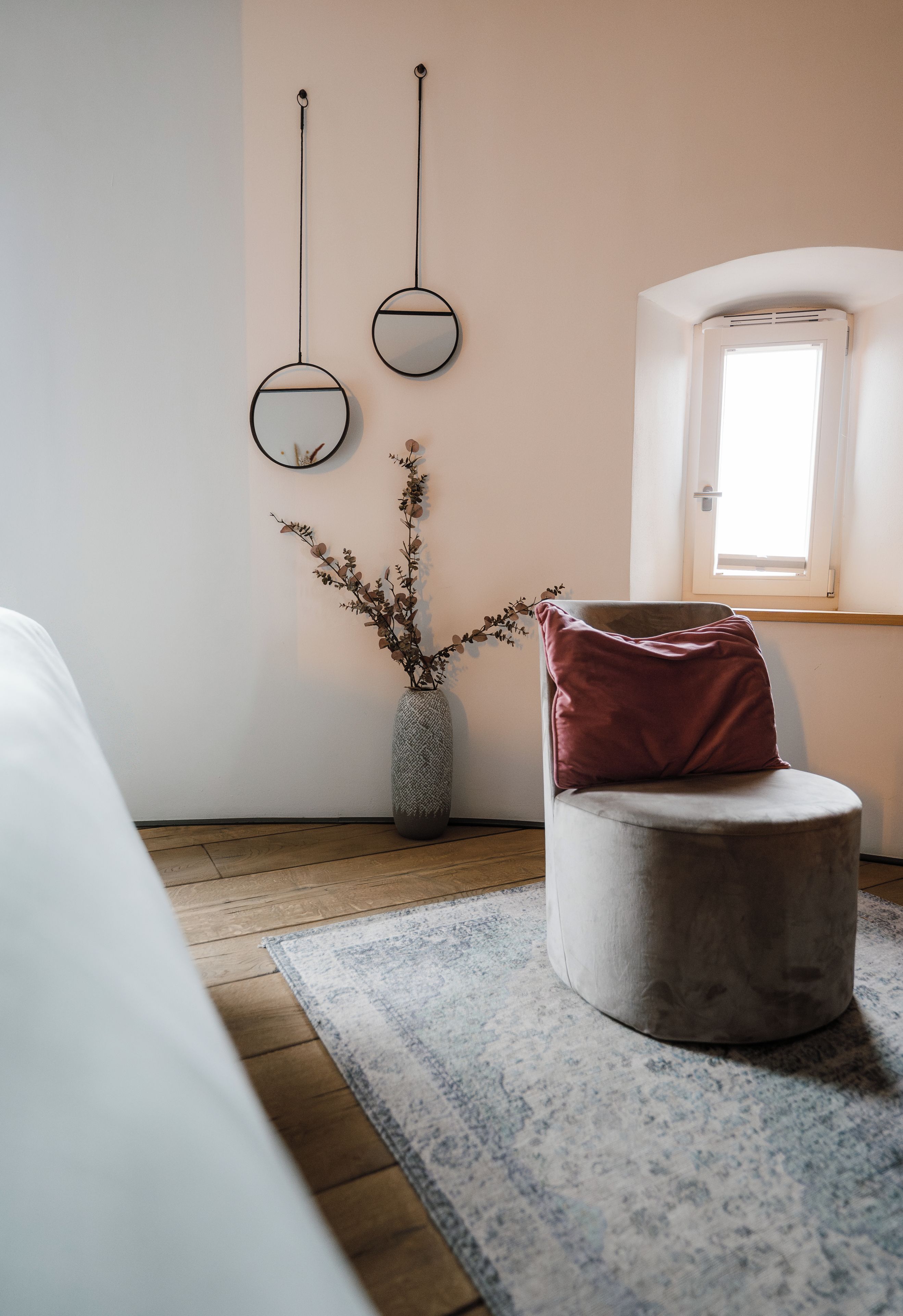 Ein modernes Zimmer mit einem runden Ottomanen, einer Vase mit Zweigen und zwei an der Wand befestigten Spiegeln.