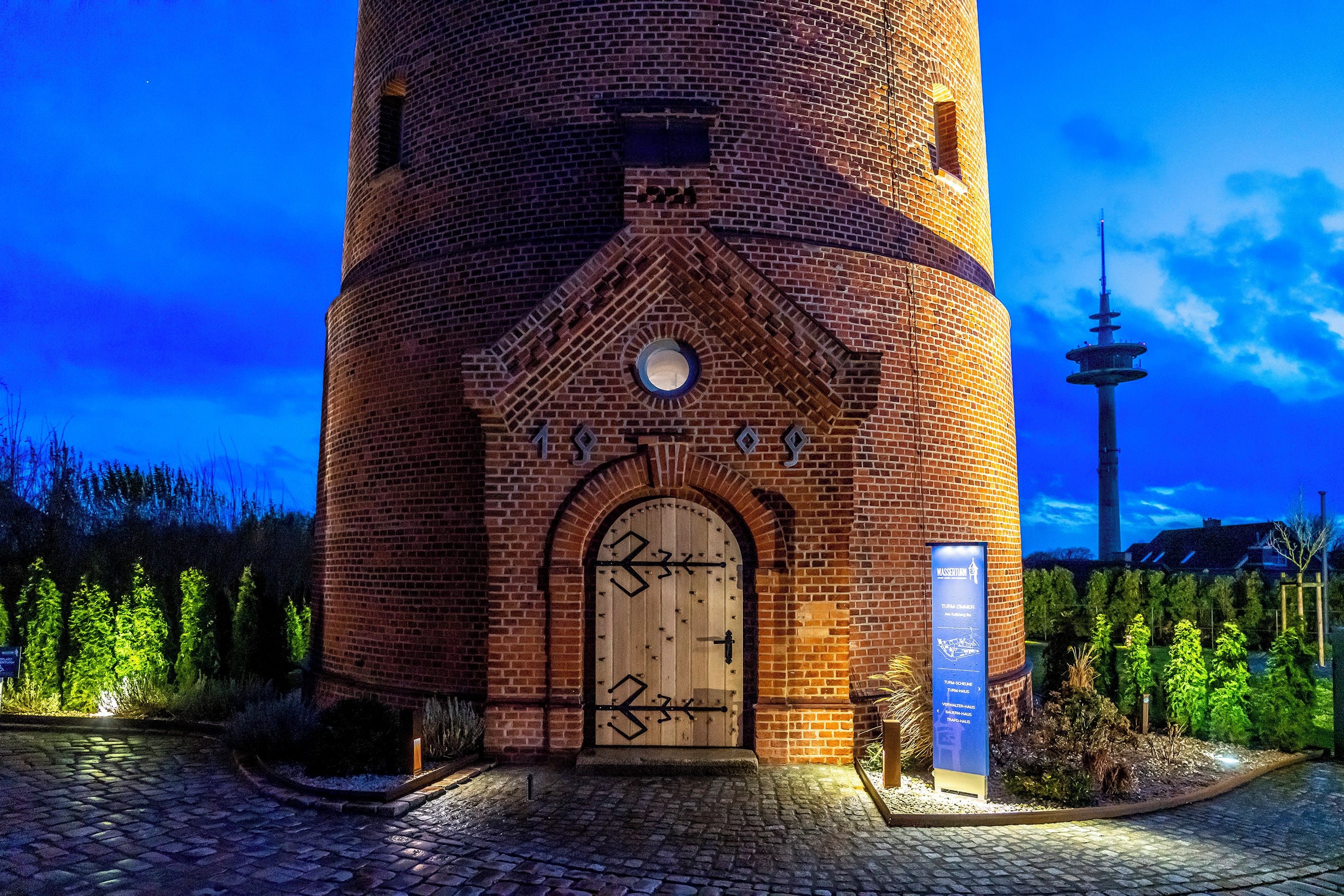 Una torre de ladrillo con una puerta de madera, un letrero y otra torre al fondo.