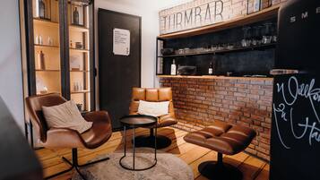 Un rincón acogedor con un sillón de cuero, una mesita redonda y un banco. Se ve una pared de ladrillo con estantes y un letrero al fondo.
