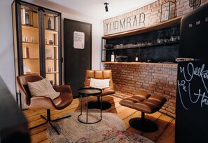 A cozy seating area with a leather chair, a small round table, and a bench. A brick wall with shelves and a sign are visible in the background. - Apart Hotel Wasserturm (Bad Segeberg)