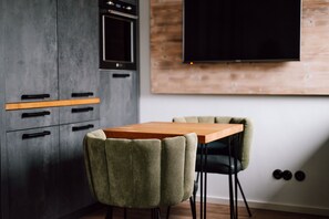 A modern kitchen with a wooden table and two chairs, a built-in oven, and a flat-screen TV mounted on the wall. - Apart Hotel Wasserturm (Bad Segeberg)