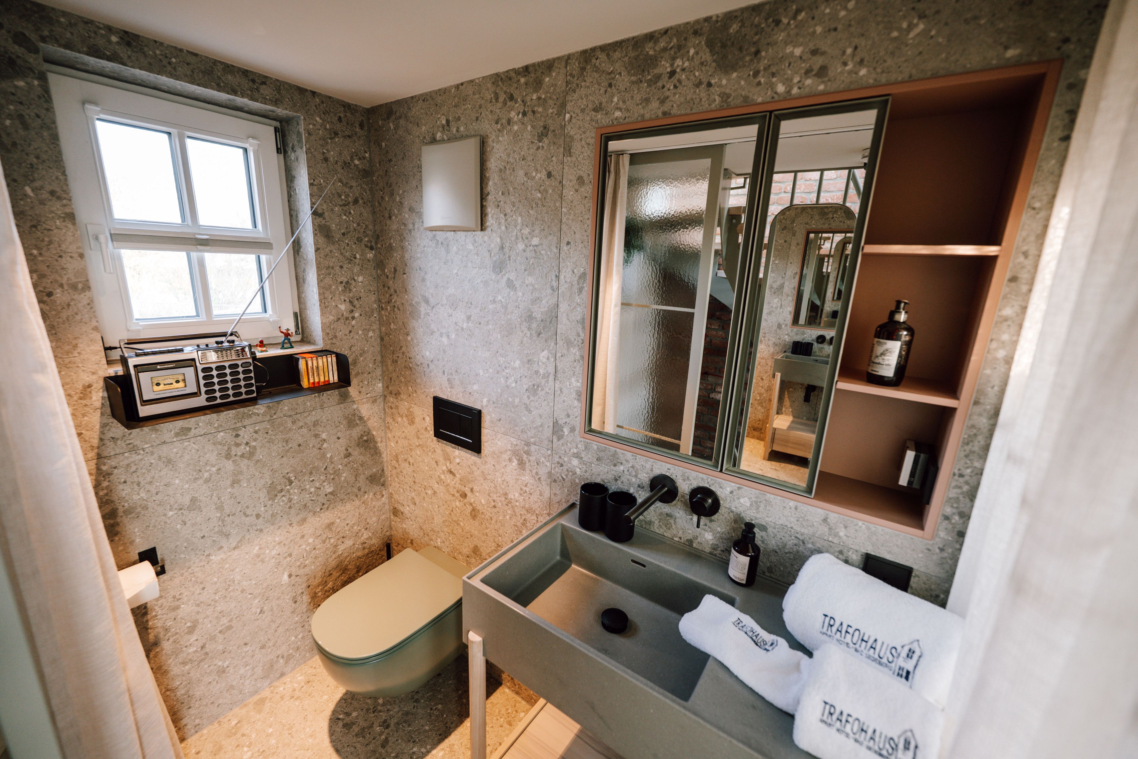 Ein kleines Badezimmer mit Toilette, Waschbecken und einem Spiegel darüber. Ein Fenster sorgt für Tageslicht und es gibt ein Ablagefach mit Pflegeprodukten.