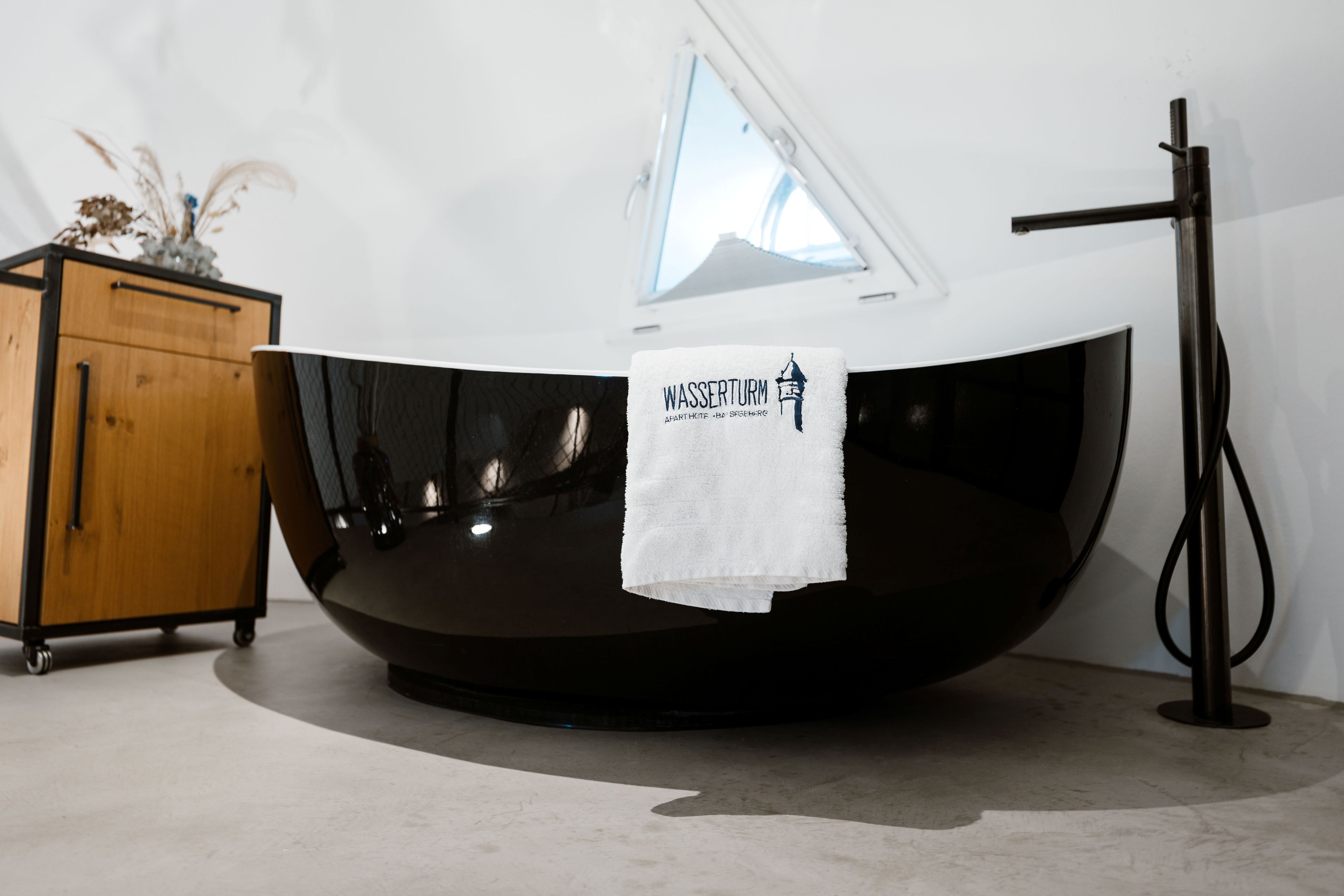 Un baño moderno con una bañera negra, un mueble de madera y una toalla con el texto 'WASSERTURM'.