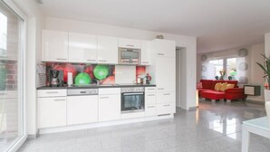 Cozinha moderna com armários brancos, forno embutido e máquina de café. Há um sofá vermelho na área de estar adjacente.