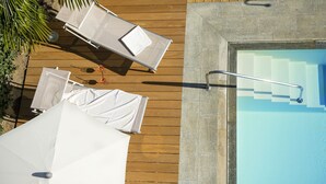 Eine Holzterrasse mit Liegestühlen und Sonnenschirmen, ein Schwimmbecken mit Metallleiter und eine Steinsäule.
