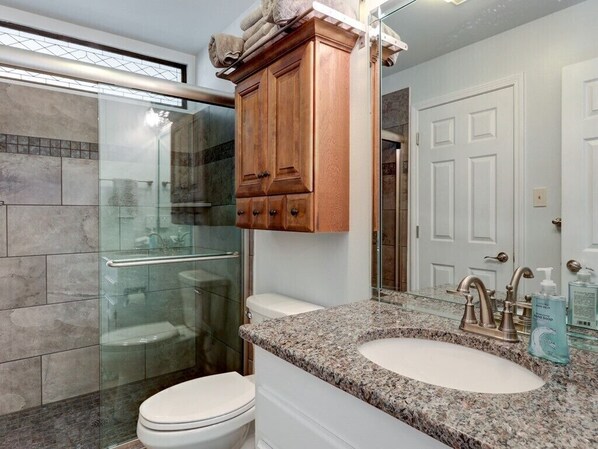Un bagno con lavabo su piano in granito, mobile bagno in legno, box doccia in vetro e WC bianco.
