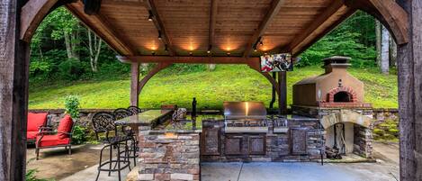 An outdoor patio with a stone barbecue and grill area, surrounded by wooden beams and a stone wall.