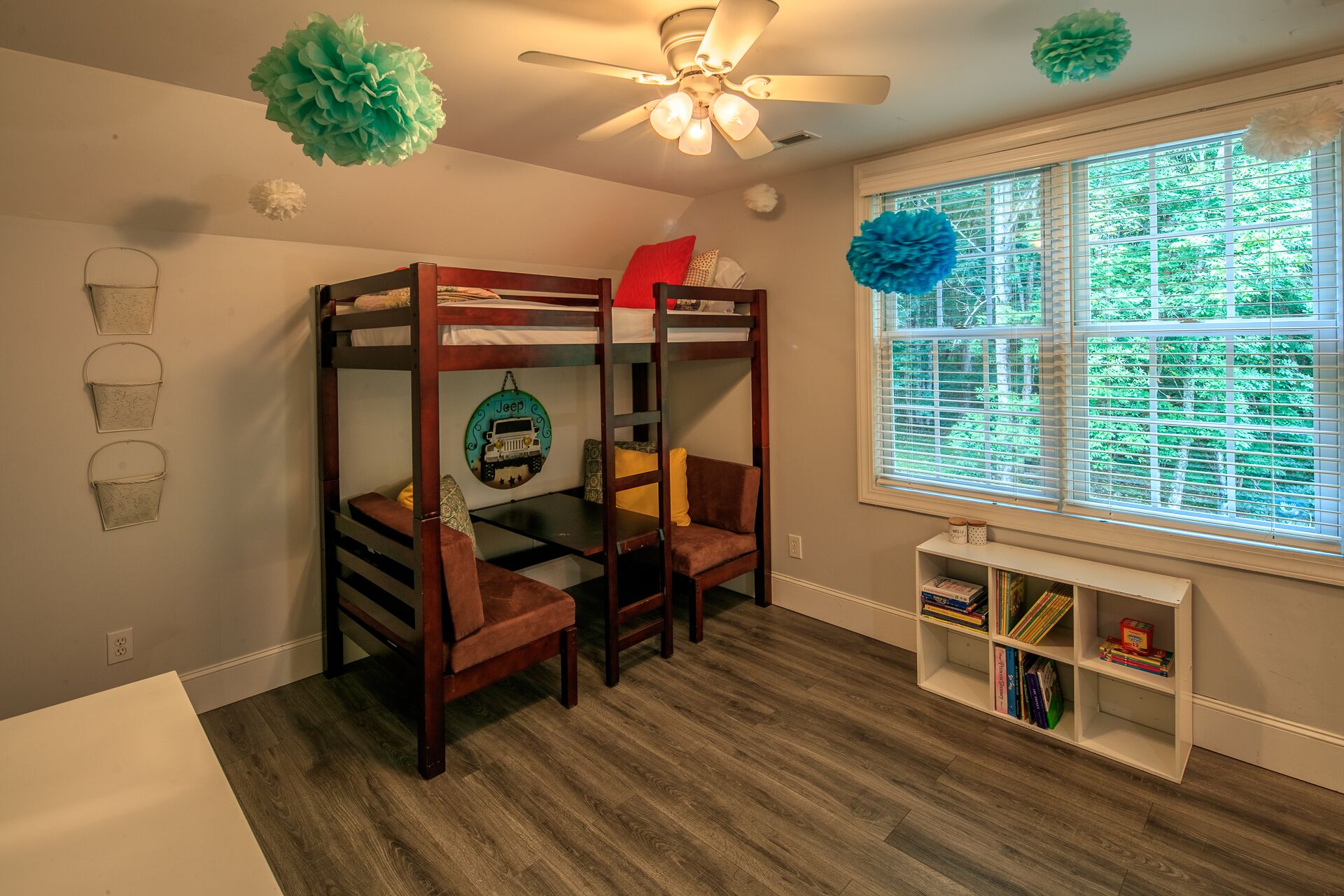 A bunk bed with a Jeep-themed wall decoration, a ceiling fan, and a window with blinds overlooking greenery.