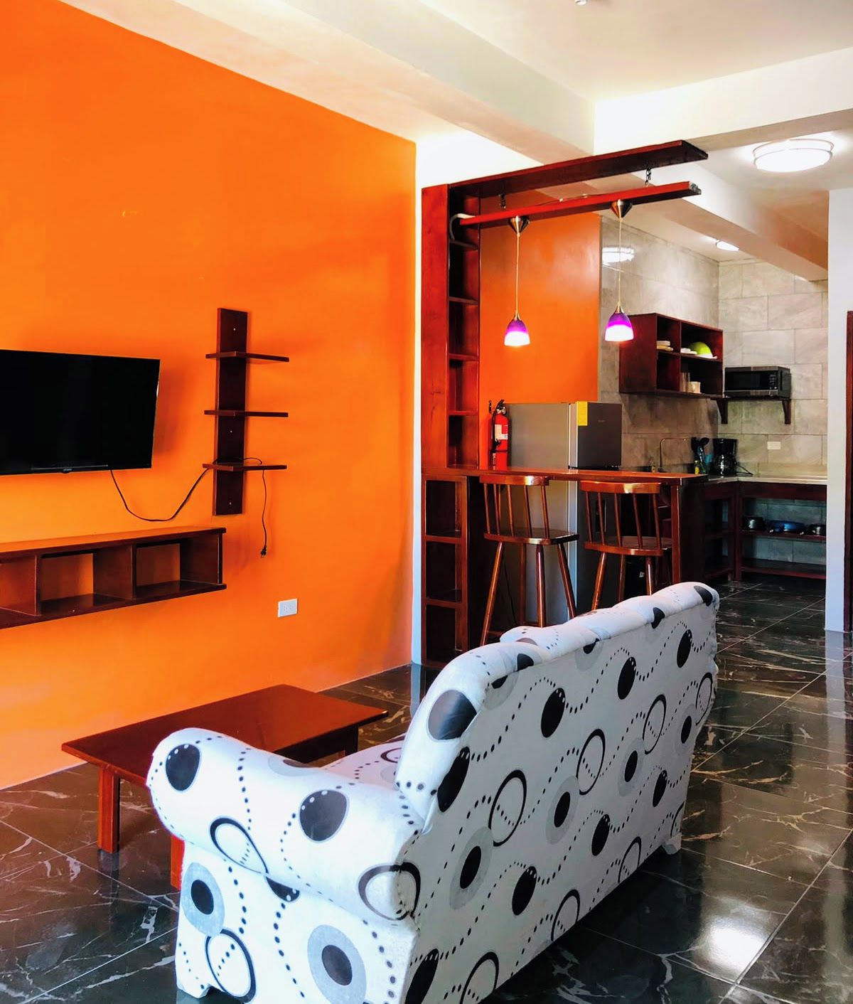 A modern living room with a sofa, a TV mounted on the wall, and a kitchen area visible through an open doorway.