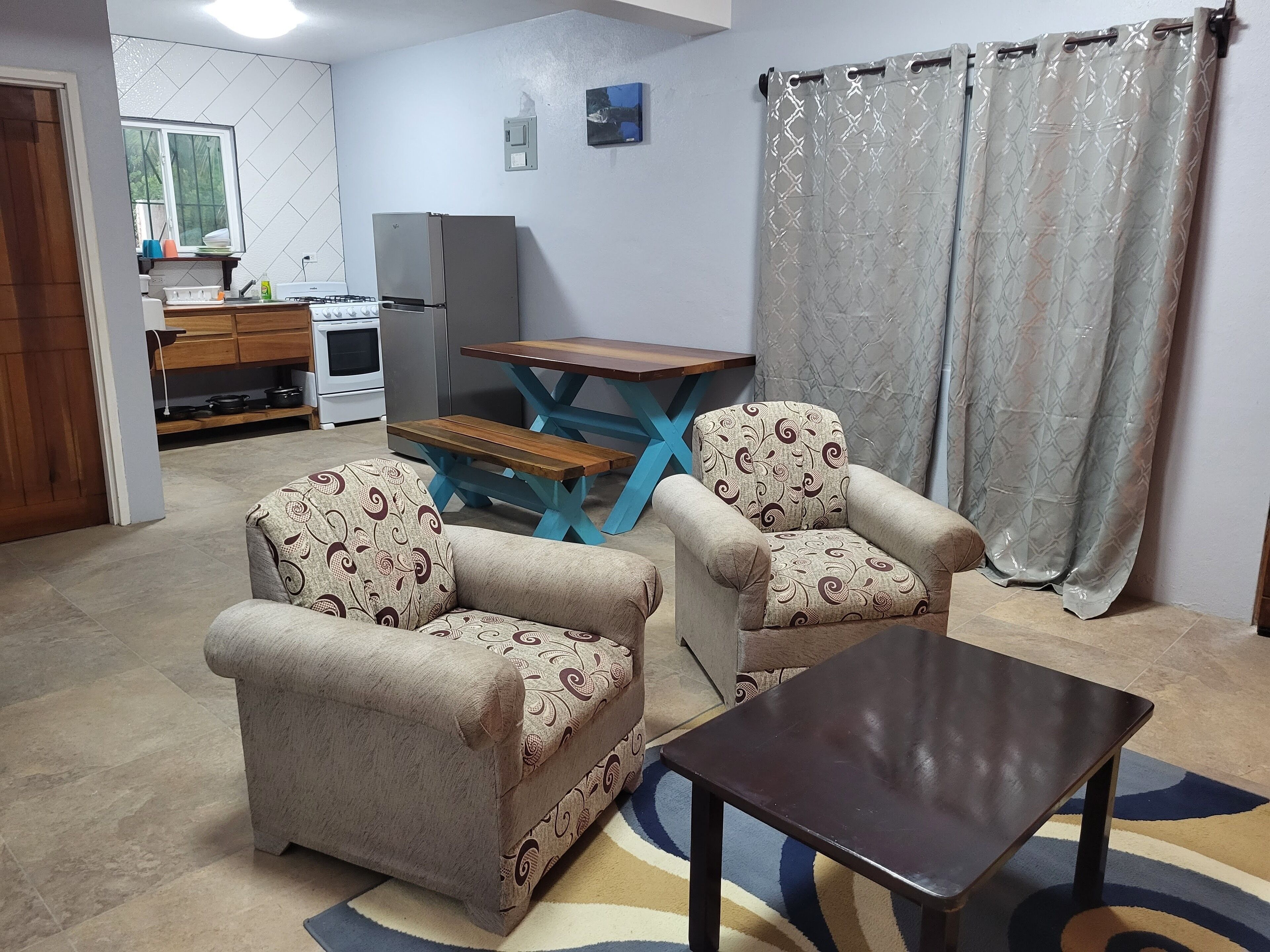 A living room with two armchairs, a wooden coffee table, a dining table with benches, a kitchen area with a refrigerator, stove, and cabinets.