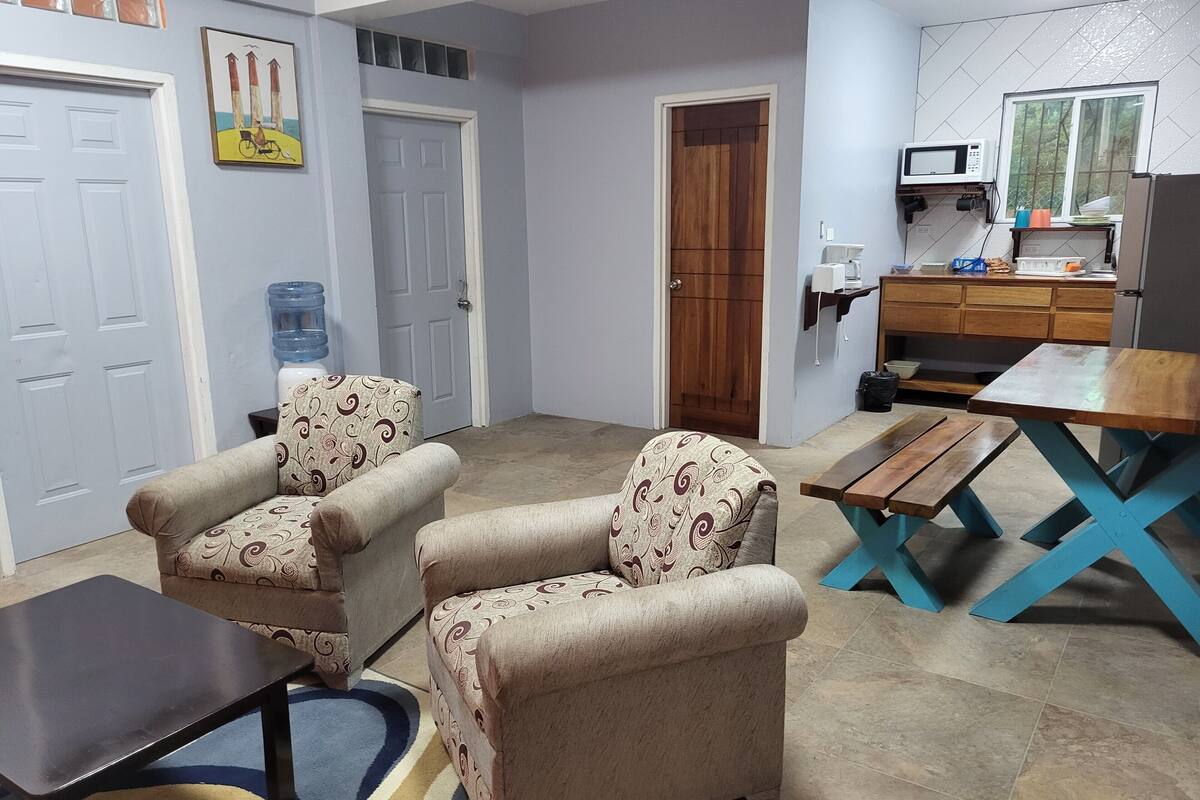 A living room with two patterned armchairs, a small table, and a wooden dining table. A kitchen area with a microwave and a water dispenser is visible in the background.