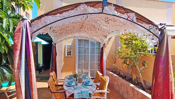 Un área de comedor al aire libre con una mesa dispuesta para cuatro personas, rodeada de sillas, bajo un arco decorativo.