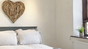 Un lit bien fait, agrémenté d’une décoration murale en forme de cœur, une fenêtre avec une plante et un radiateur.