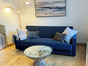 Um sofá azul escuro com duas almofadas, uma mesa de centro de vidro com design exclusivo e uma pintura na parede.