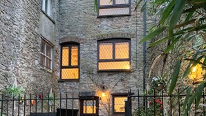 Un bâtiment en pierre doté de nombreuses fenêtres, dont certaines sont équipées de moustiquaires, et d’une petite porte. On aperçoit une clôture en métal au premier plan, ainsi que quelques plantes.