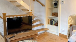Un salon avec un téléviseur à écran plat posé sur un meuble en bois, une table ronde en pierre, un escalier en bois, une bibliothèque et un mur en pierre.
