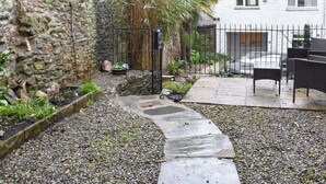 Une cour avec un chemin en pierre, une clôture en métal noir et un espace détente aménagé avec des meubles en osier.