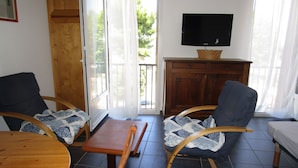 Sala de estar com duas poltronas de balanço, uma mesa de madeira, uma TV em um móvel e uma porta de vidro corrediça que dá para uma varanda.