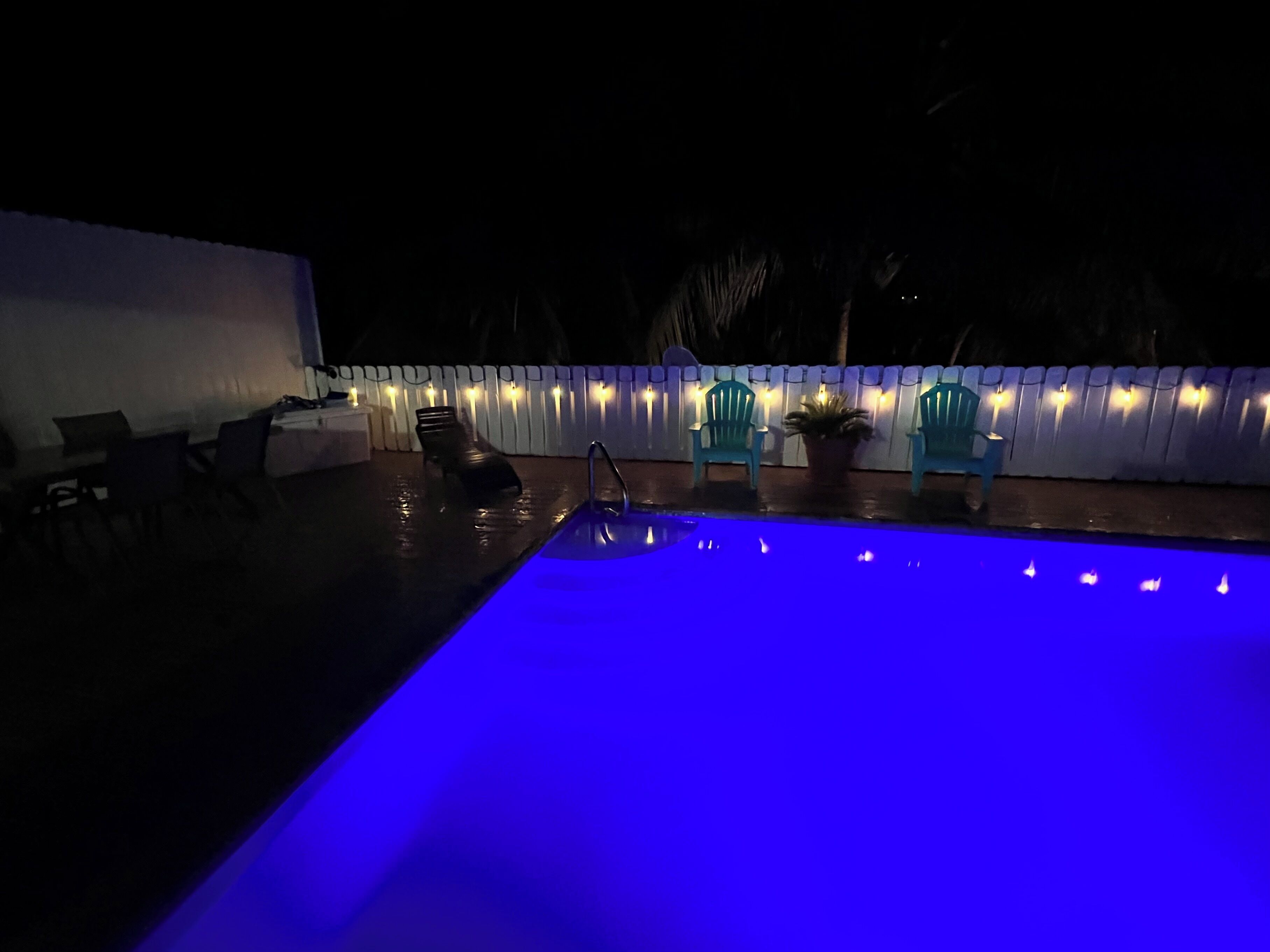 Une piscine extérieure dont le bord est illuminé de lumière bleue, entourée de chaises et d’une clôture blanche décorée de guirlandes lumineuses.
