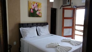 A neatly made bed with white linens and two pillows, a painting of pink flowers on the wall, and a window with a wooden frame. - Hotel Praia da Paixão (Prado)