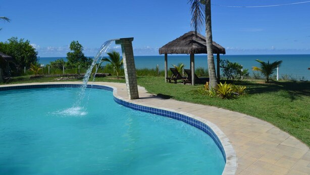 A swimming pool with a clear blue water feature, a thatched gazebo, and a view of the ocean. - Hotel Praia da Paixão (Prado)