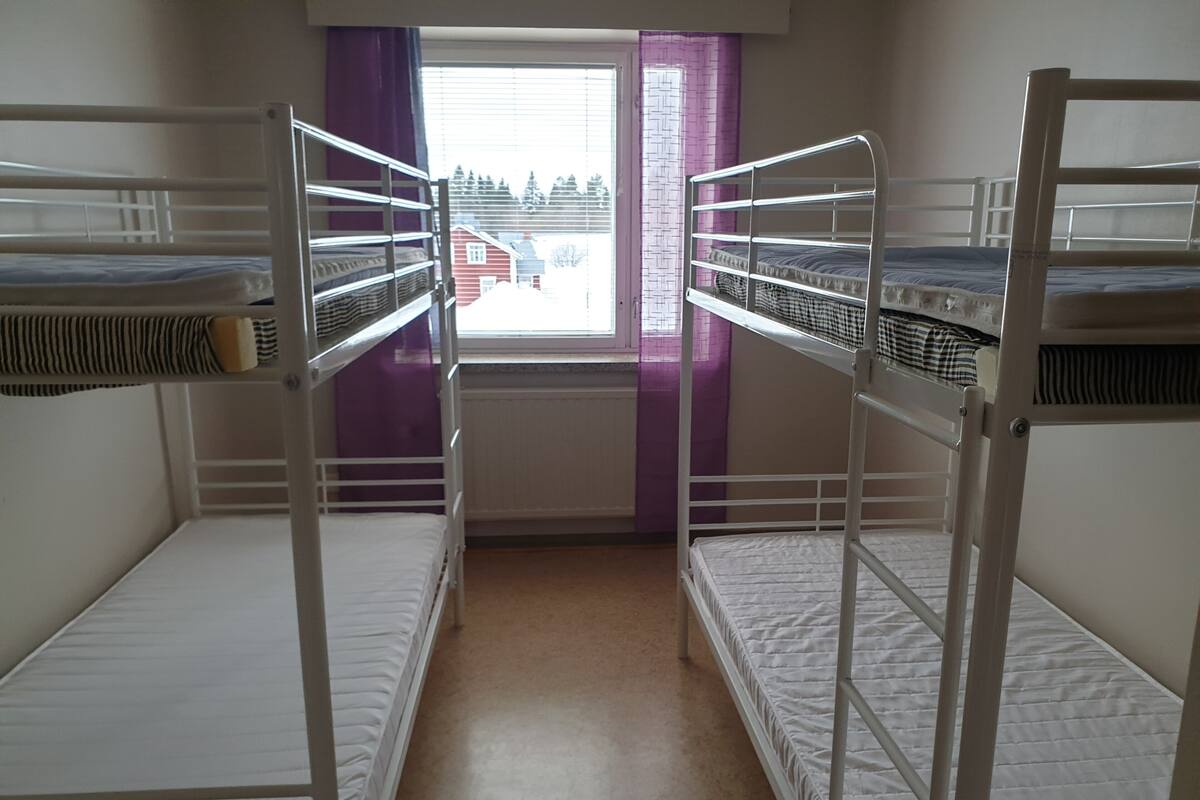 A dormitory room with bunk beds, a window with purple curtains, and a view of houses outside.