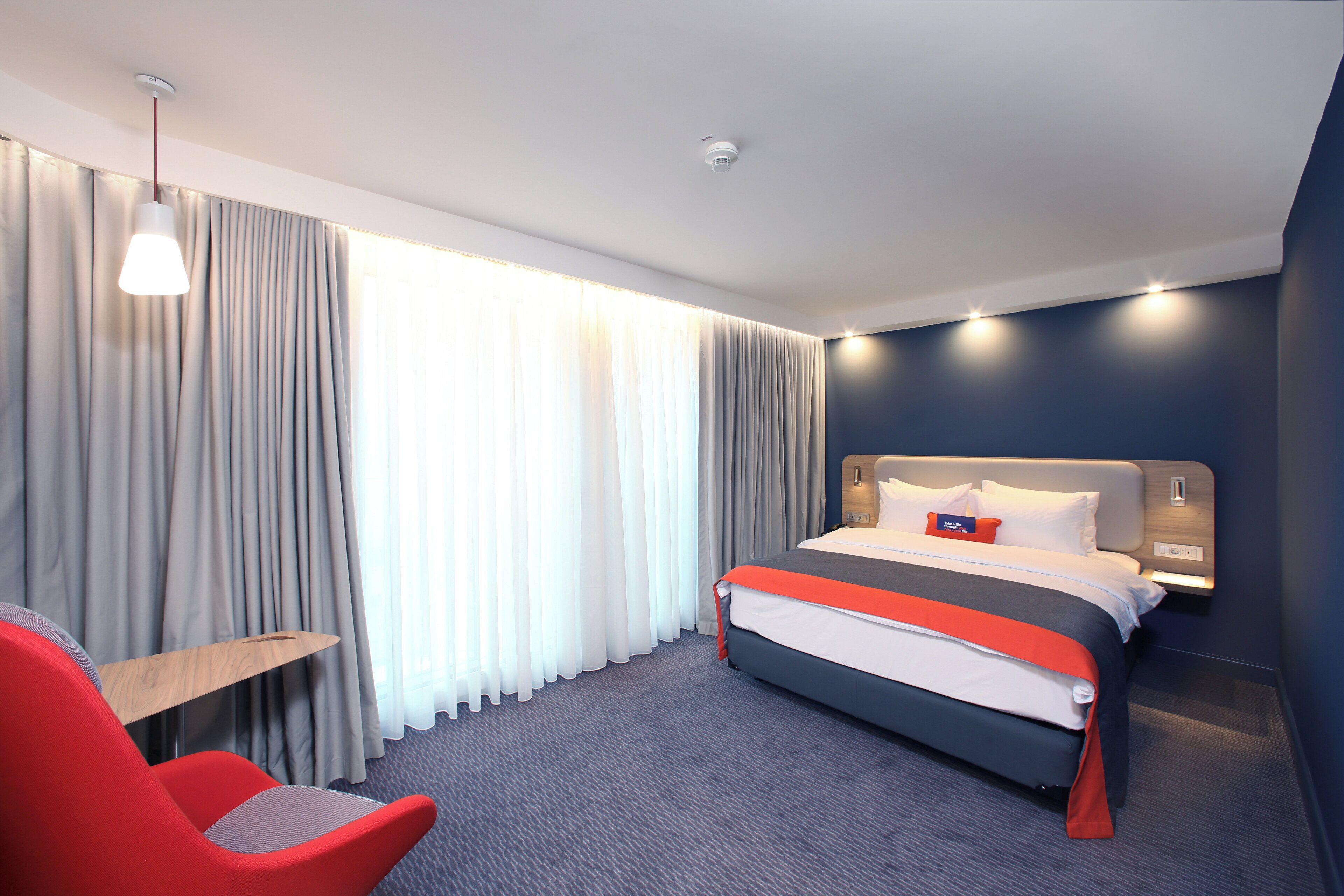 Una habitación de hotel con cama, escritorio, silla y un gran ventanal con cortinas.