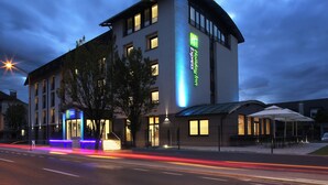 Un hôtel moderne, reconnaissable à son grand panneau vert, doté d’un espace extérieur abrité avec des sièges et d’une entrée bien éclairée.