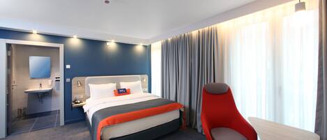 Habitación de hotel moderna con cama, escritorio, silla, baño y un gran ventanal con cortinas.