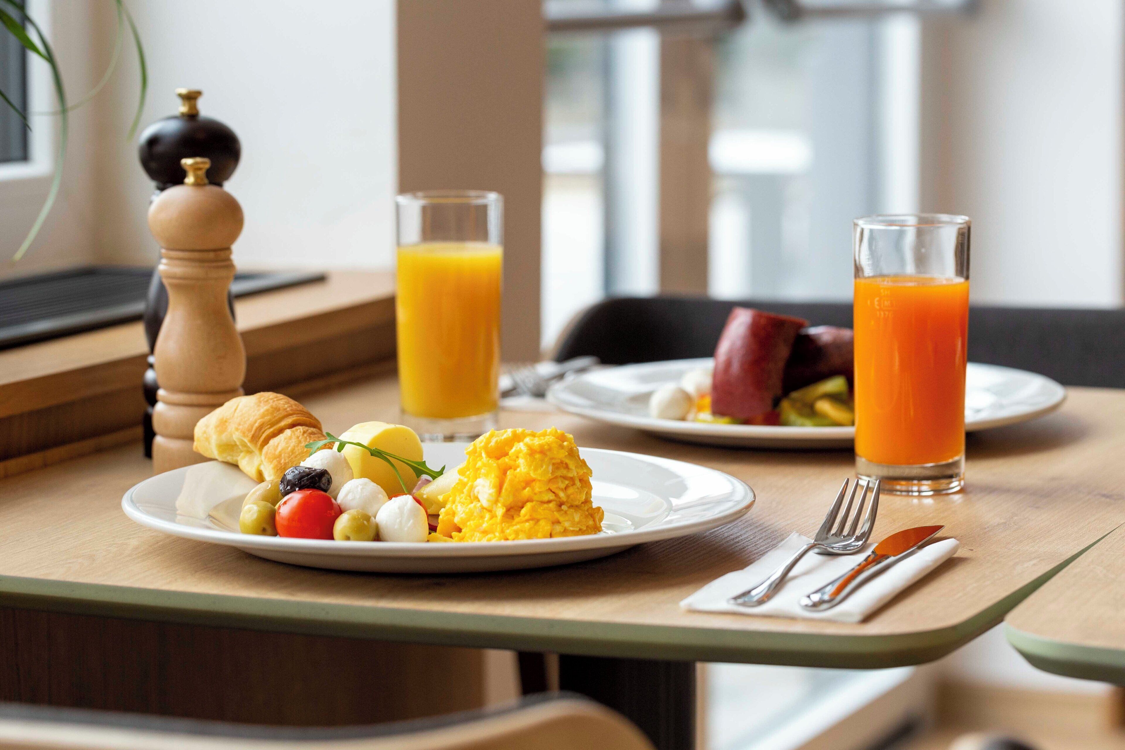 Une table dressée pour le petit-déjeuner avec des assiettes contenant des œufs brouillés, des tomates, des olives et un croissant, ainsi que deux verres de jus d’orange.
