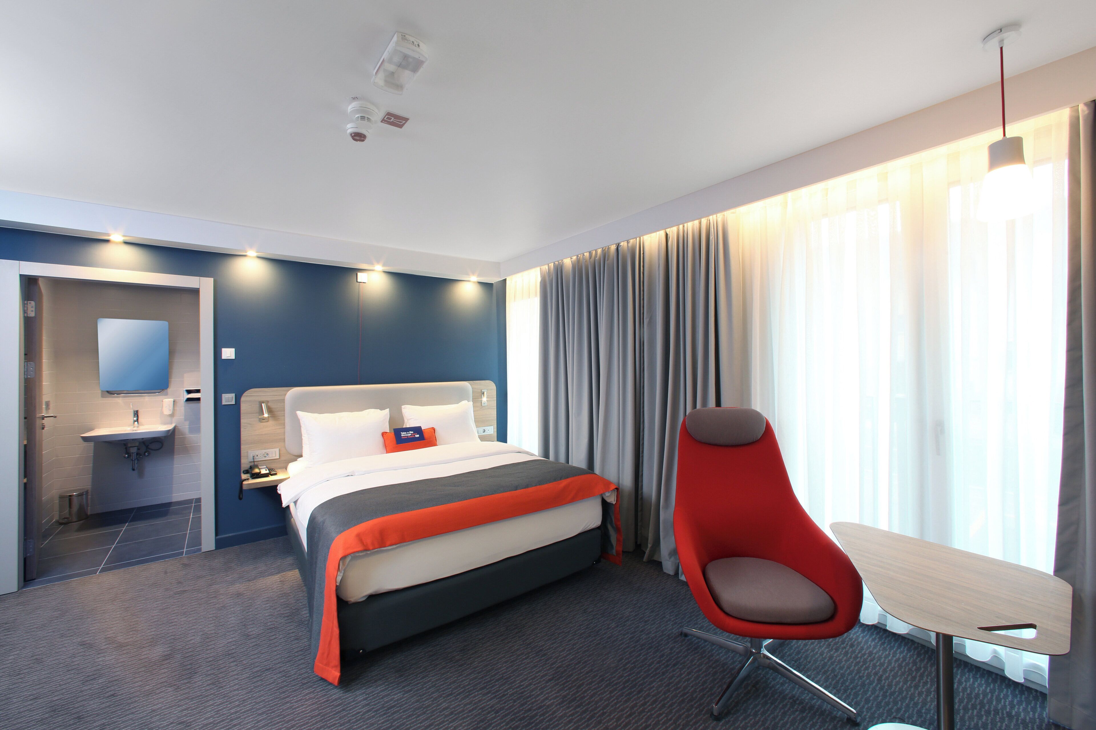 Habitación de hotel con cama, escritorio, silla, lámpara de escritorio, espejo y ventana con cortinas. Incluye baño con lavabo y espejo.
