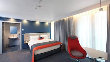 Habitación de hotel con cama, escritorio, silla, lámpara de escritorio, espejo y ventana con cortinas. Incluye baño con lavabo y espejo.