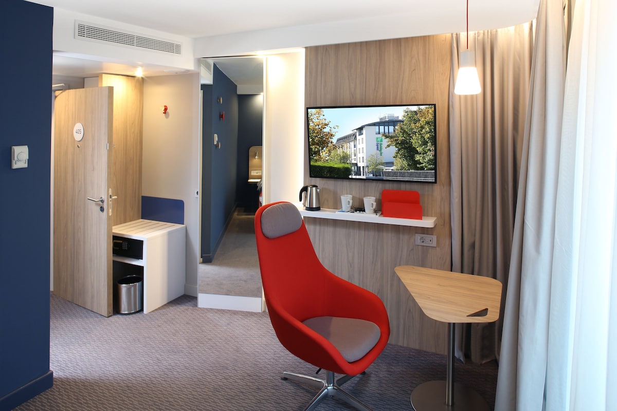 Ett modernt hotellrum med en röd stol, ett litet bord, en tv som är monterad på väggen och utsikt över staden genom fönstret.