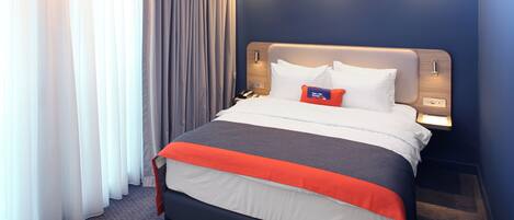 A hotel room with a bed, curtains, a nightstand, and a wall-mounted lamp.