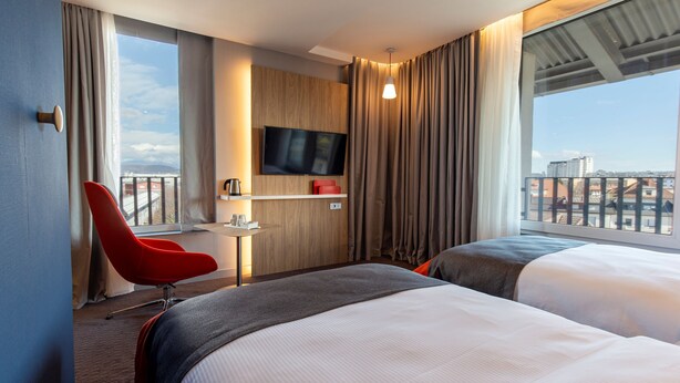 Une chambre d’hôtel avec deux lits, un fauteuil rouge, un téléviseur fixé au mur et une vue sur la ville.