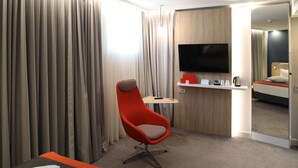 Une chambre d’hôtel dotée d’un grand téléviseur à écran plat, d’un fauteuil rouge, d’une petite table avec une télécommande et d’un lit agrémenté d’une touche de couleur orange.