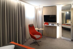 Habitación de hotel con un televisor de pantalla plana grande, un sillón rojo, una mesita con un control remoto y una cama con detalles naranja.