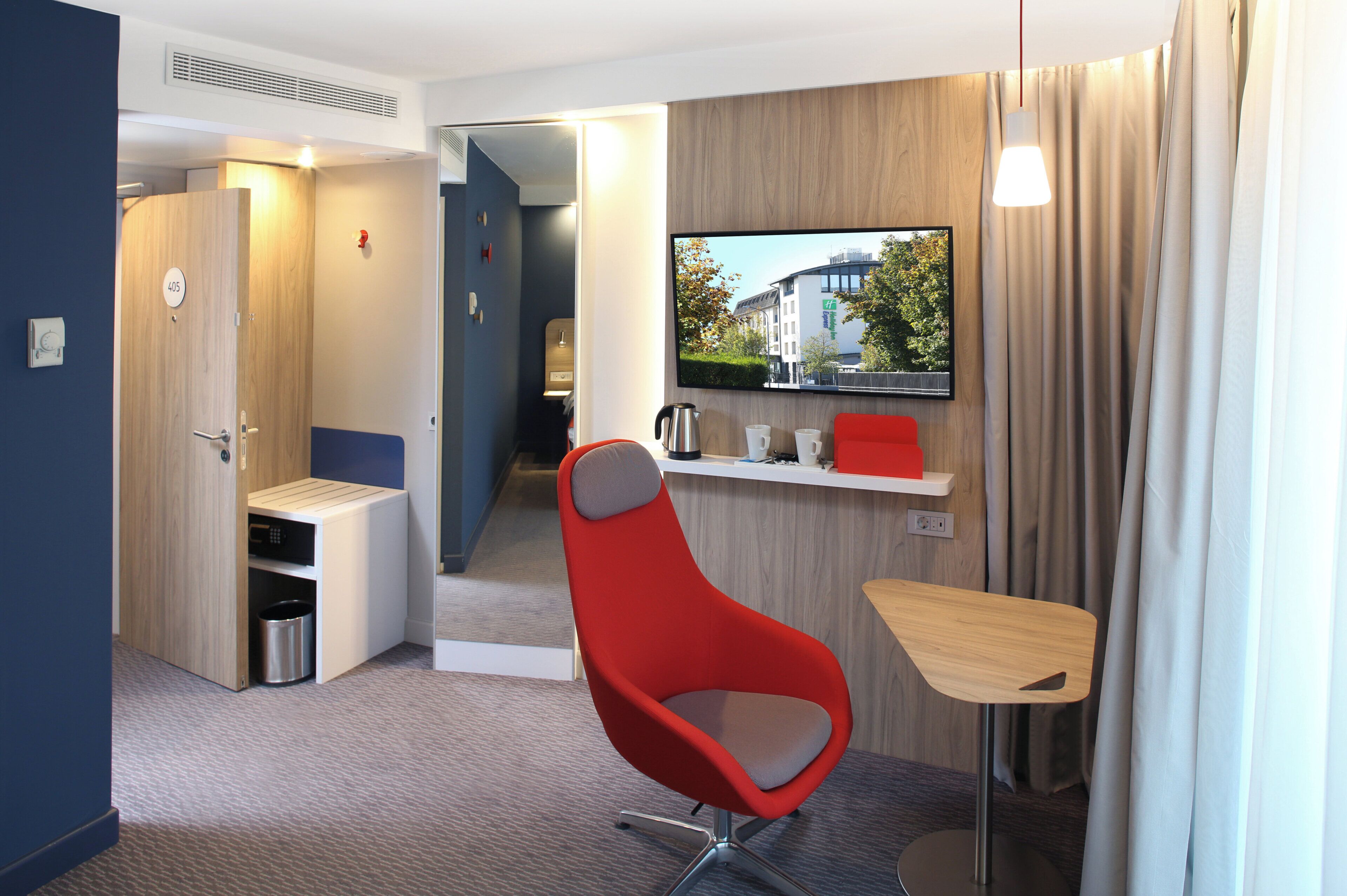 Ein modernes Hotelzimmer mit einem roten Sessel, einem kleinen Tisch, einem an der Wand montierten Fernseher und Blick ins Freie.