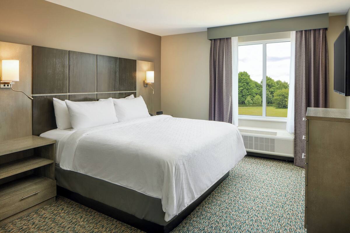 A hotel room with a large bed, a nightstand, a desk, a flat-screen TV, and a window with curtains.