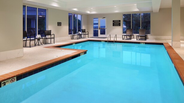Una amplia piscina interior con agua cristalina y azul, rodeada de ventanas y sillas.